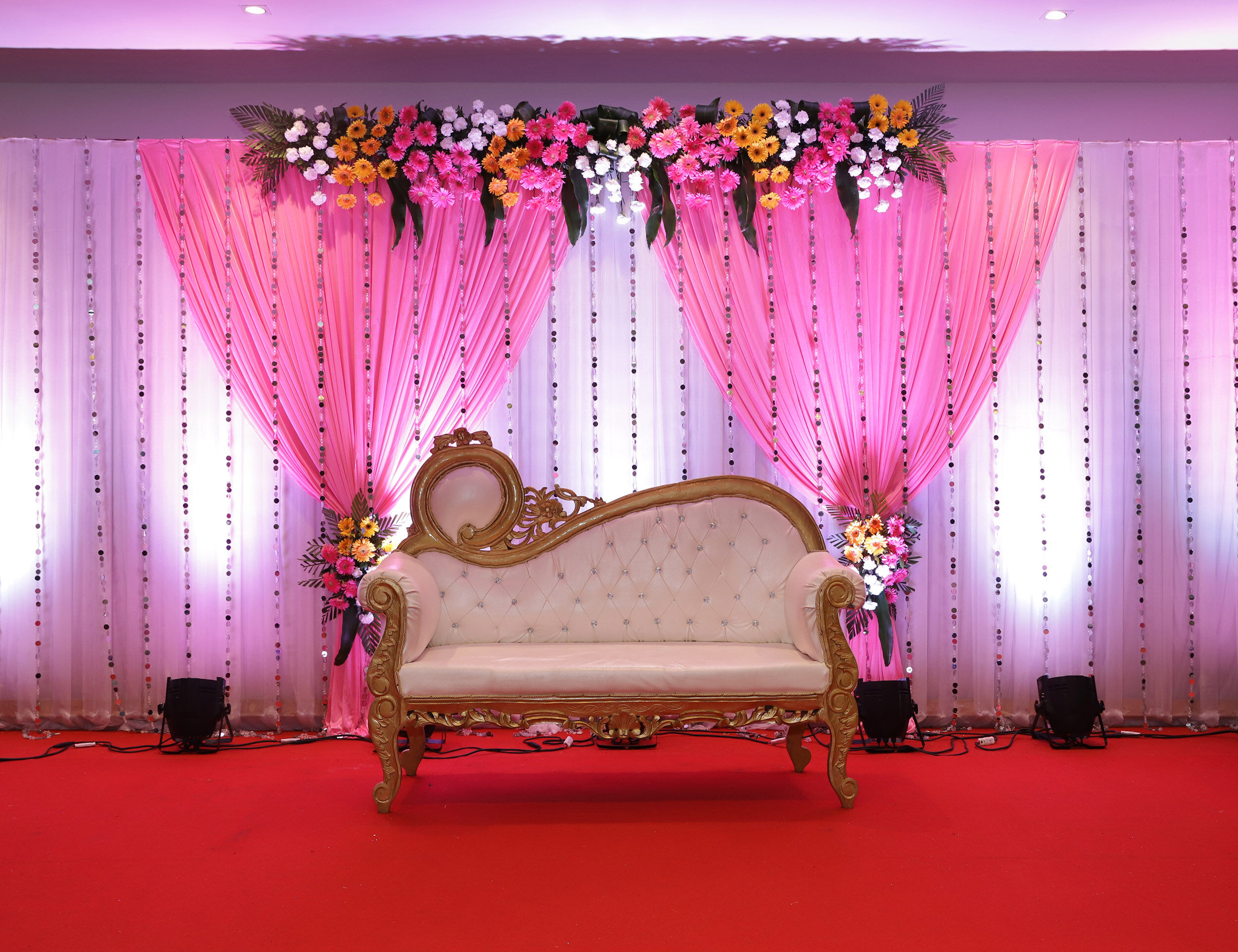 Pink-themed indoor wedding stage with floral arch, elegant Victorian-style sofa, and draped curtains on red carpet